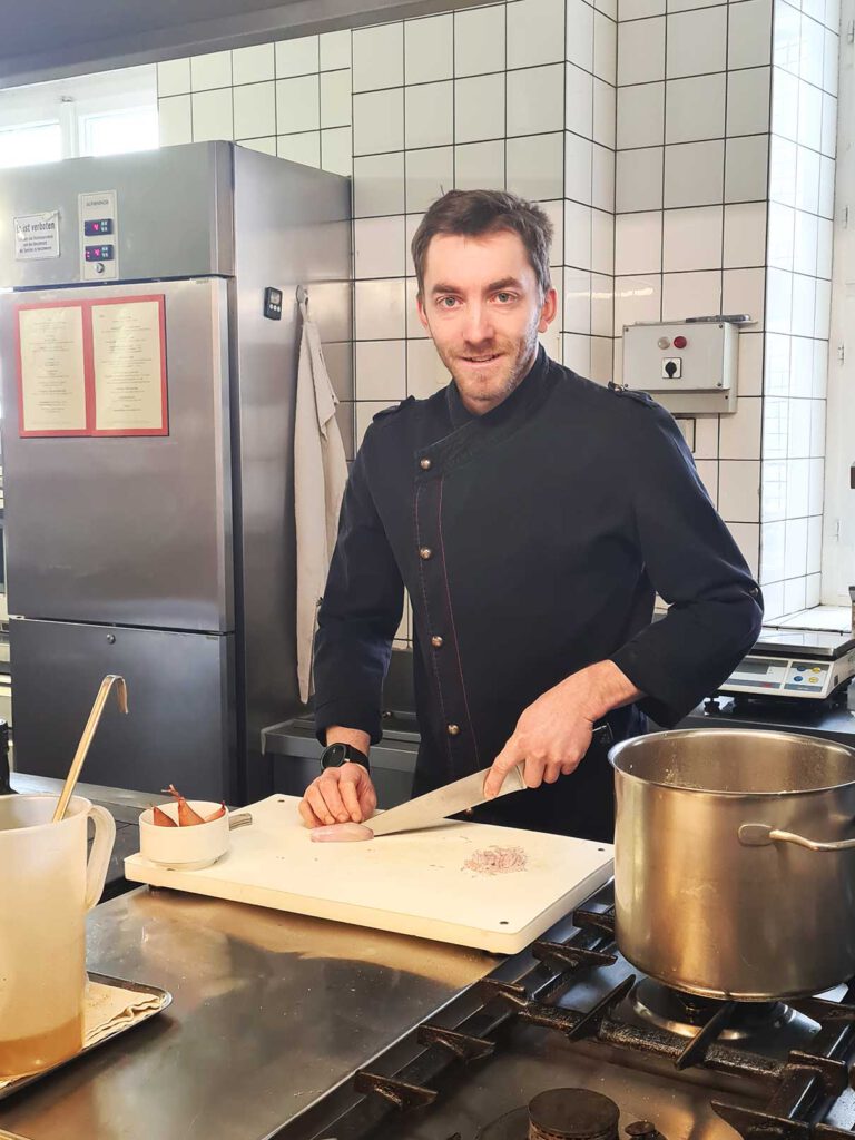 Ein Koch in schwarzer Uniform lächelt, während er in einer professionellen Küche Zutaten auf einem Schneidebrett zerkleinert.